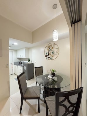 A modern dining area with a glass table, two chairs, a decorative wall piece, and a kitchen in the background. - COZI Uptown Parksuites (Taguig)