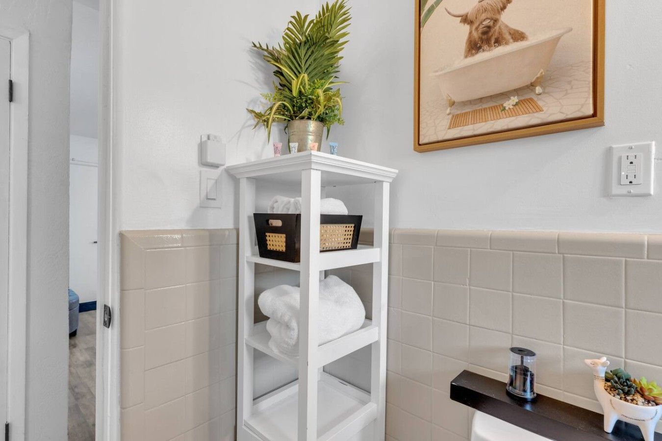 A bathroom with a white shelf holding towels and a potted plant, a framed picture on the wall, and a small plant on the counter.