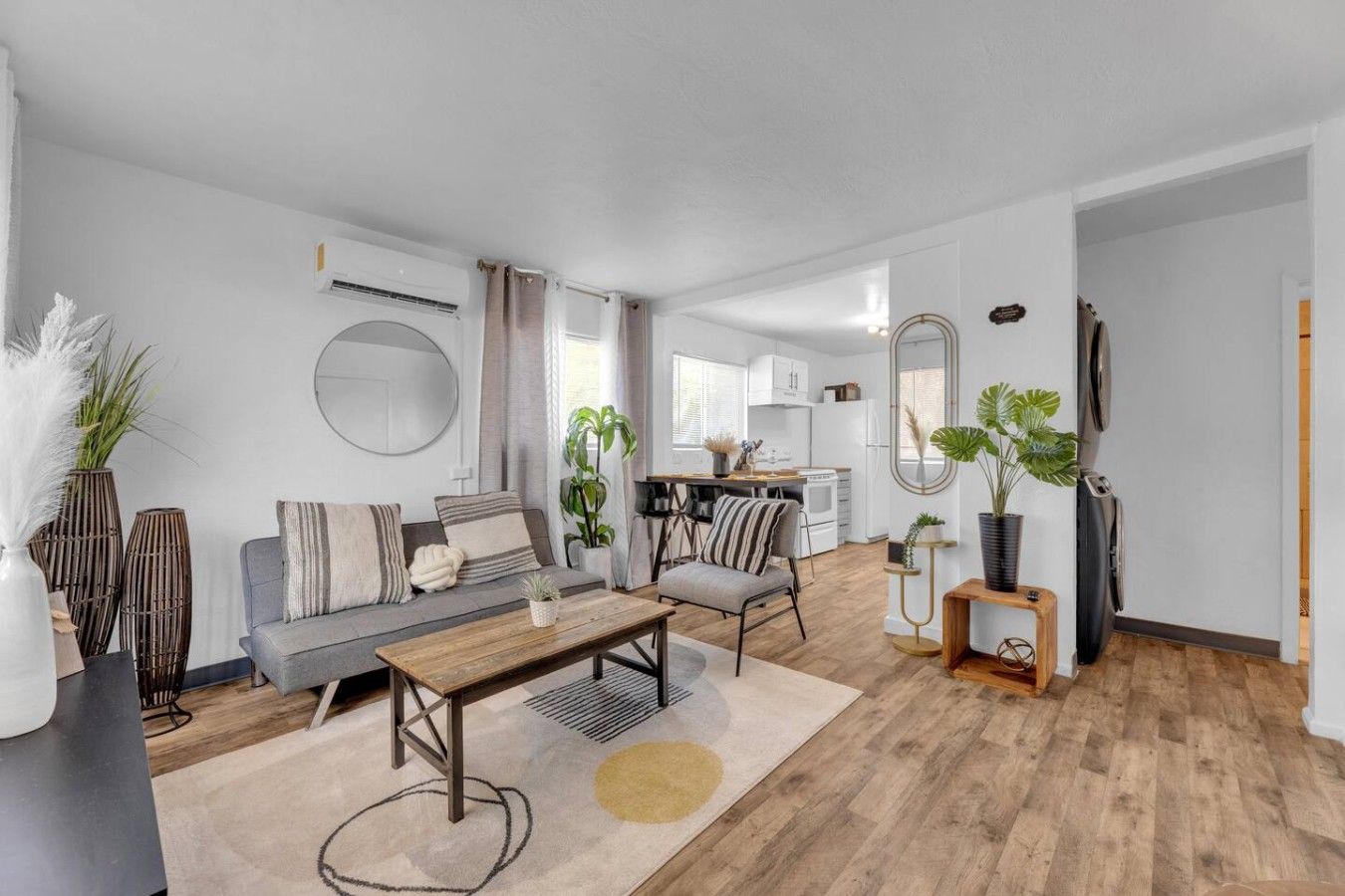 A modern living room with a sofa, coffee table, and chairs, a kitchen area with cabinets and a dining table, and a hallway with a mirror and plants.