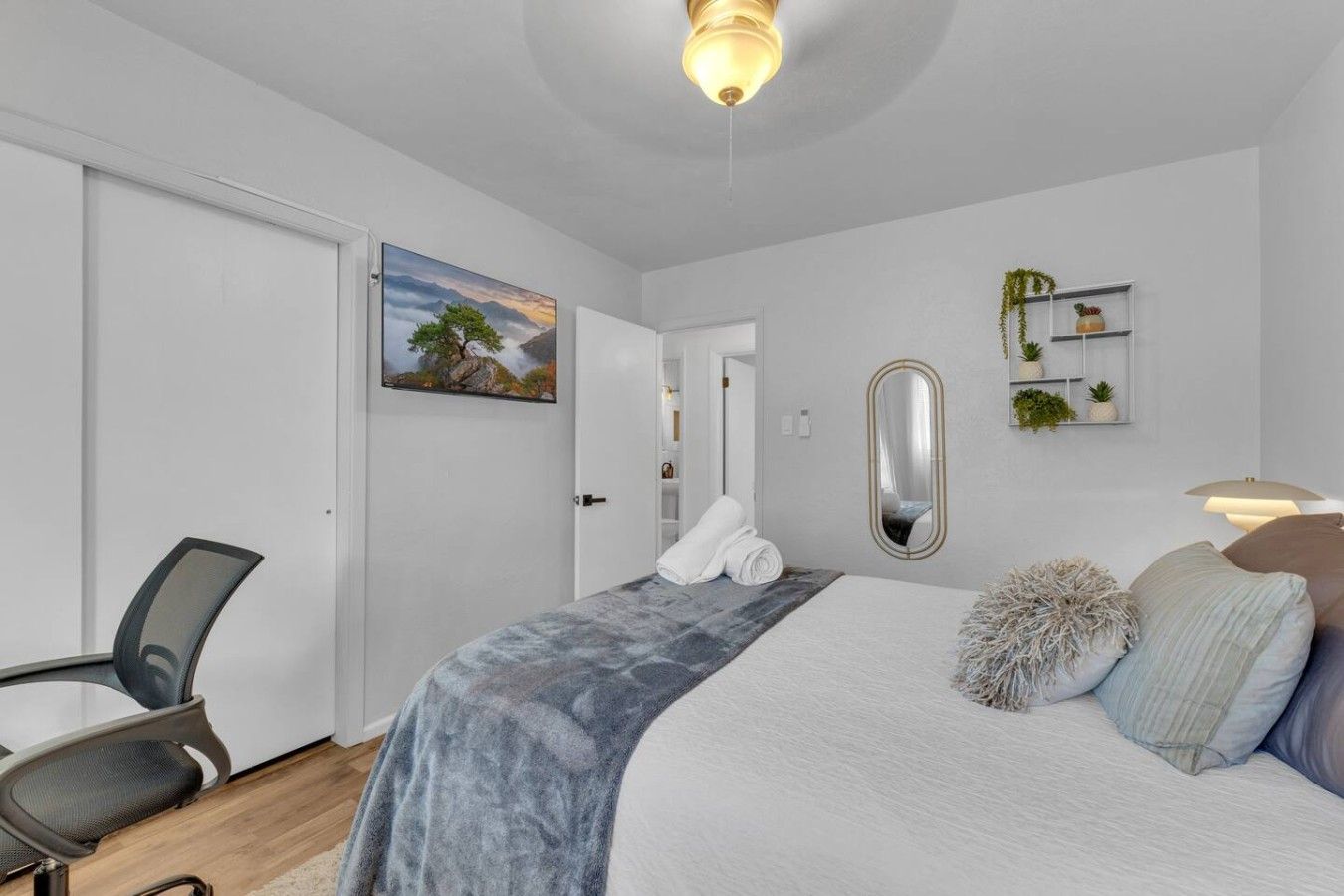 Een slaapkamer met een bed, een stoel, een bureau, een schilderij, een spiegel en een plank met planten.