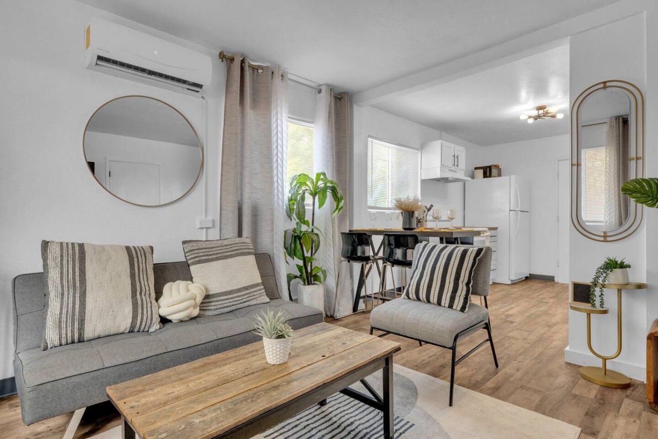 A modern apartment with a grey sofa, a wooden coffee table, a dining area with chairs, a kitchen with white cabinets, and a large mirror.
