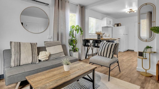 A modern apartment with a grey sofa, a wooden coffee table, a dining area with chairs, a kitchen with white cabinets, and a large mirror. - Central 2BR | Workspace, Laundry & Long Stays (El Paso)