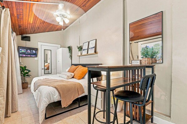 A bedroom with a bed, a desk, two chairs, a TV, and a window with blinds.