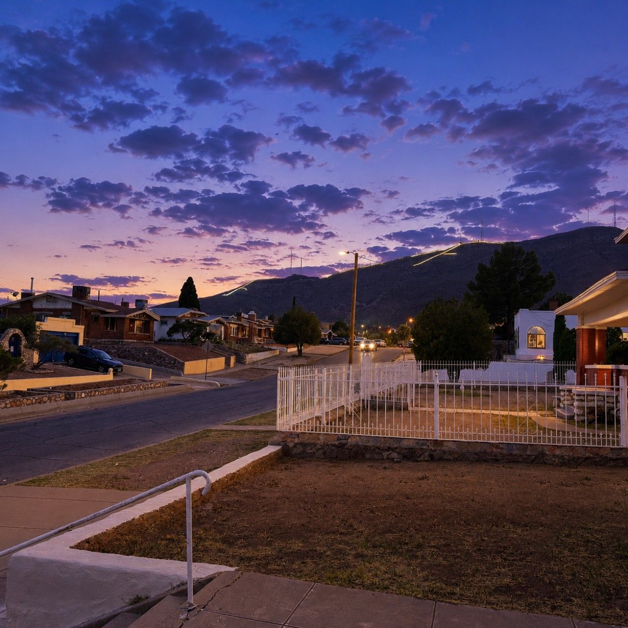 Une rue résidentielle au crépuscule, avec des maisons, une zone délimitée par une clôture et une montagne en arrière-plan.