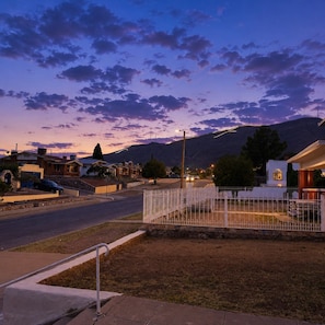 Une rue résidentielle au crépuscule, avec des maisons, une zone délimitée par une clôture et une montagne en arrière-plan.