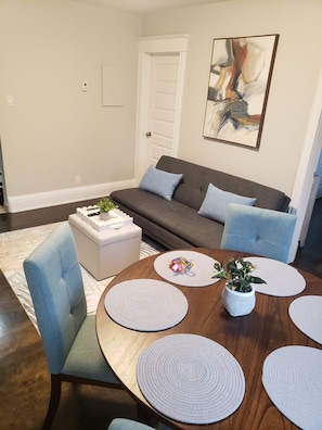 A living room with a dark grey sofa, light blue cushions, a white coffee table, and a round wooden dining table with placemats. - "Courtyard Arms"  family friendly (Toronto)