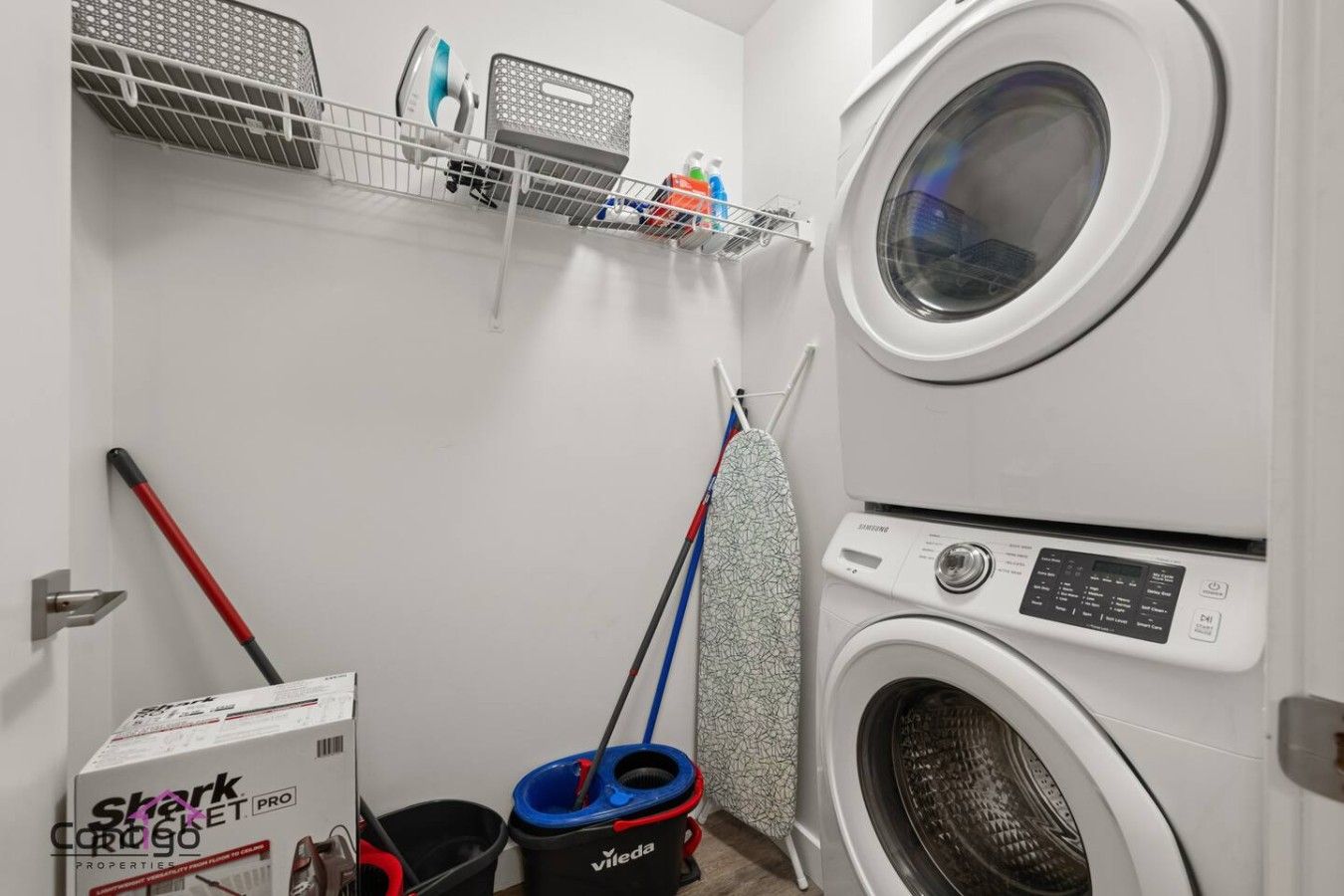 A laundry room with a washing machine, a mopping bucket, a mop, and a box labeled 'Shark Cortez PRO'.