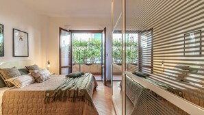 Une chambre à coucher avec un lit, des oreillers, une lampe de chevet, une fenêtre avec des stores et une vue sur la verdure à l’extérieur.