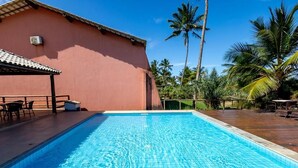 Piscina com deck ladrilhado, área de estar coberta e palmeiras ao fundo.