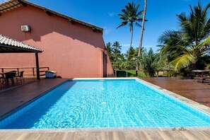 Una piscina con plataforma de baldosas, zona de estar techada y palmeras al fondo.