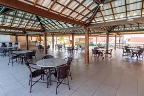 Un patio al aire libre con mesas redondas, sillas de mimbre, una zona de estar cubierta y una cocina visible al fondo.