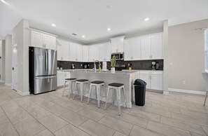 A modern kitchen with white cabinets, a stainless steel refrigerator, a central island with bar stools, and a black trash bin. - Low-Tox Theme Park Retreat • 2 Private Theaters (Four Corners)