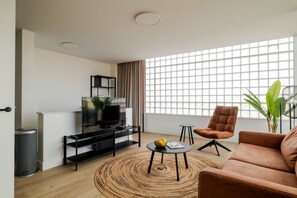 Ein modernes Wohnzimmer mit einer Couch, einem Fernseher auf einem schwarzen Schrank, einem runden Couchtisch und einem großen Fenster mit Blick.