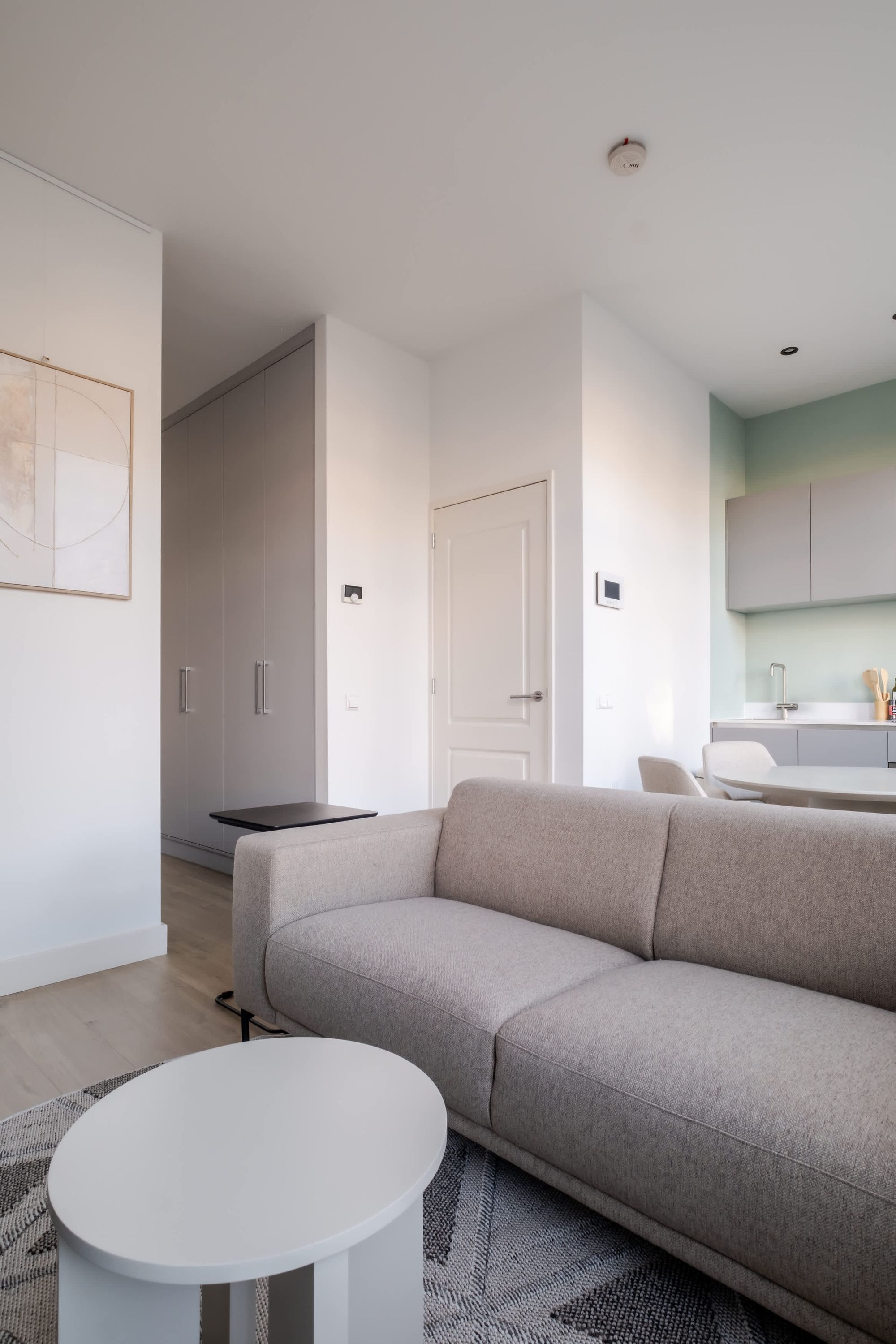 Un salon moderne avec un canapé gris, une table basse blanche et un coin cuisine doté d’un plan de travail et de meubles de rangement blancs.