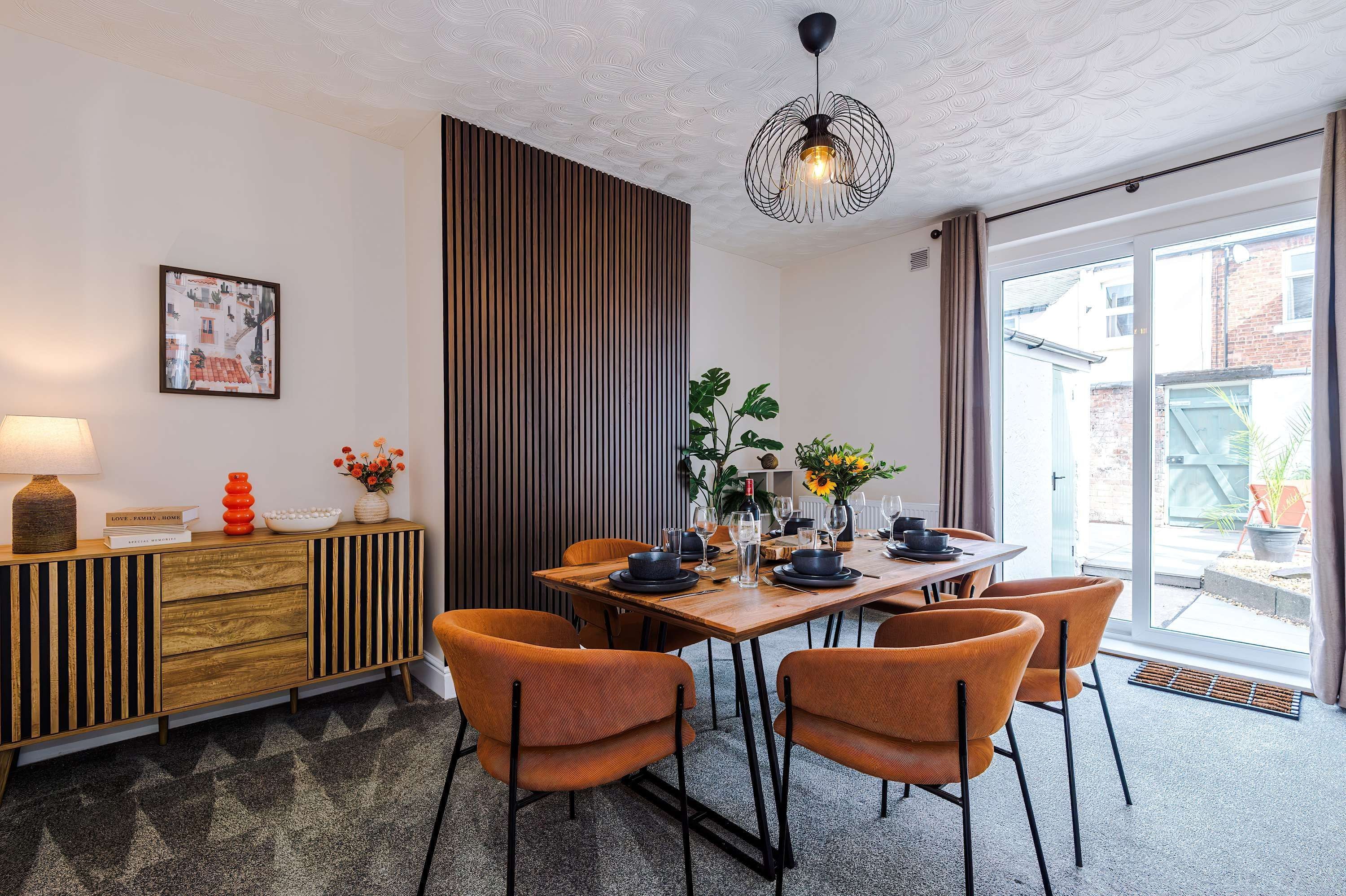 Een eetruimte met een houten tafel voor vier personen, omringd door oranje stoelen, een moderne hanglamp en een aan de muur bevestigd schilderij.