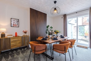 Een eetruimte met een houten tafel voor vier personen, omringd door oranje stoelen, een moderne hanglamp en een aan de muur bevestigd schilderij.
