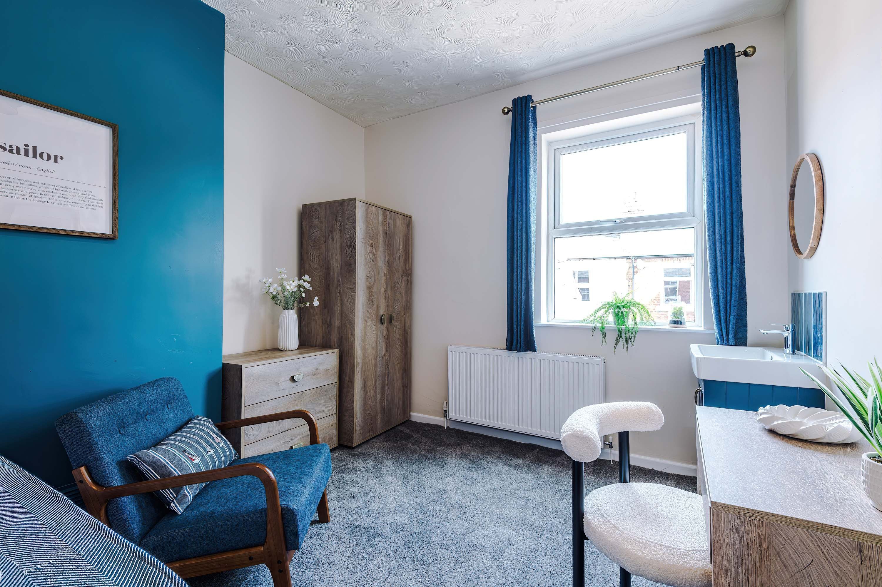 Een kamer met een blauwe wand, een houten garderobe, een radiator, een raam met blauwe gordijnen, een witte radiator, een wastafel en een stoel met een witte kussen.