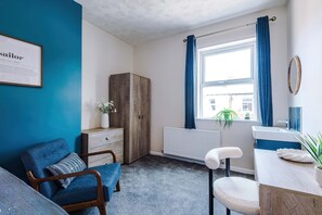 Een kamer met een blauwe wand, een houten garderobe, een radiator, een raam met blauwe gordijnen, een witte radiator, een wastafel en een stoel met een witte kussen.