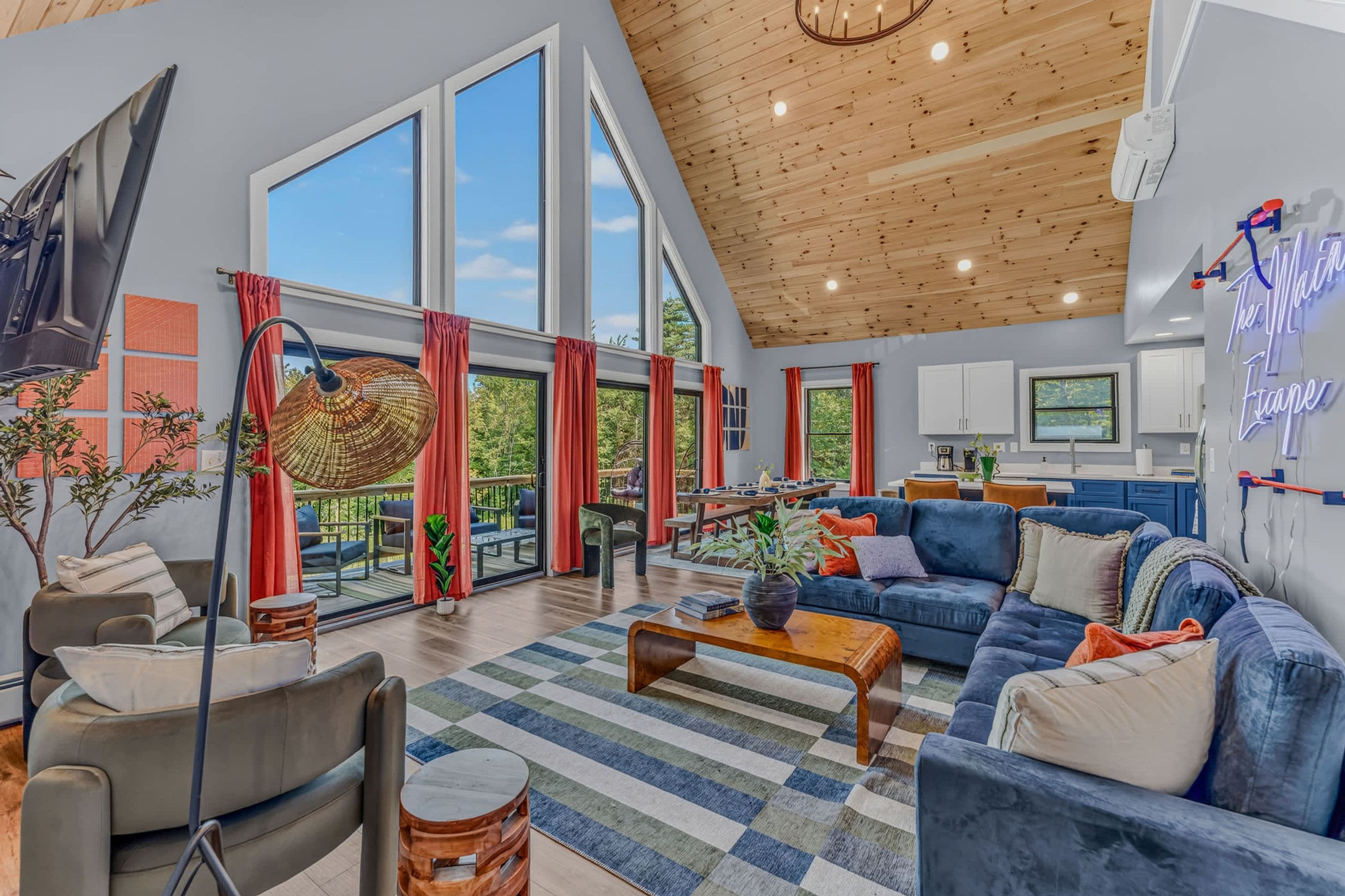 A spacious living area with large windows, a wooden ceiling, and a modern kitchen in the background.