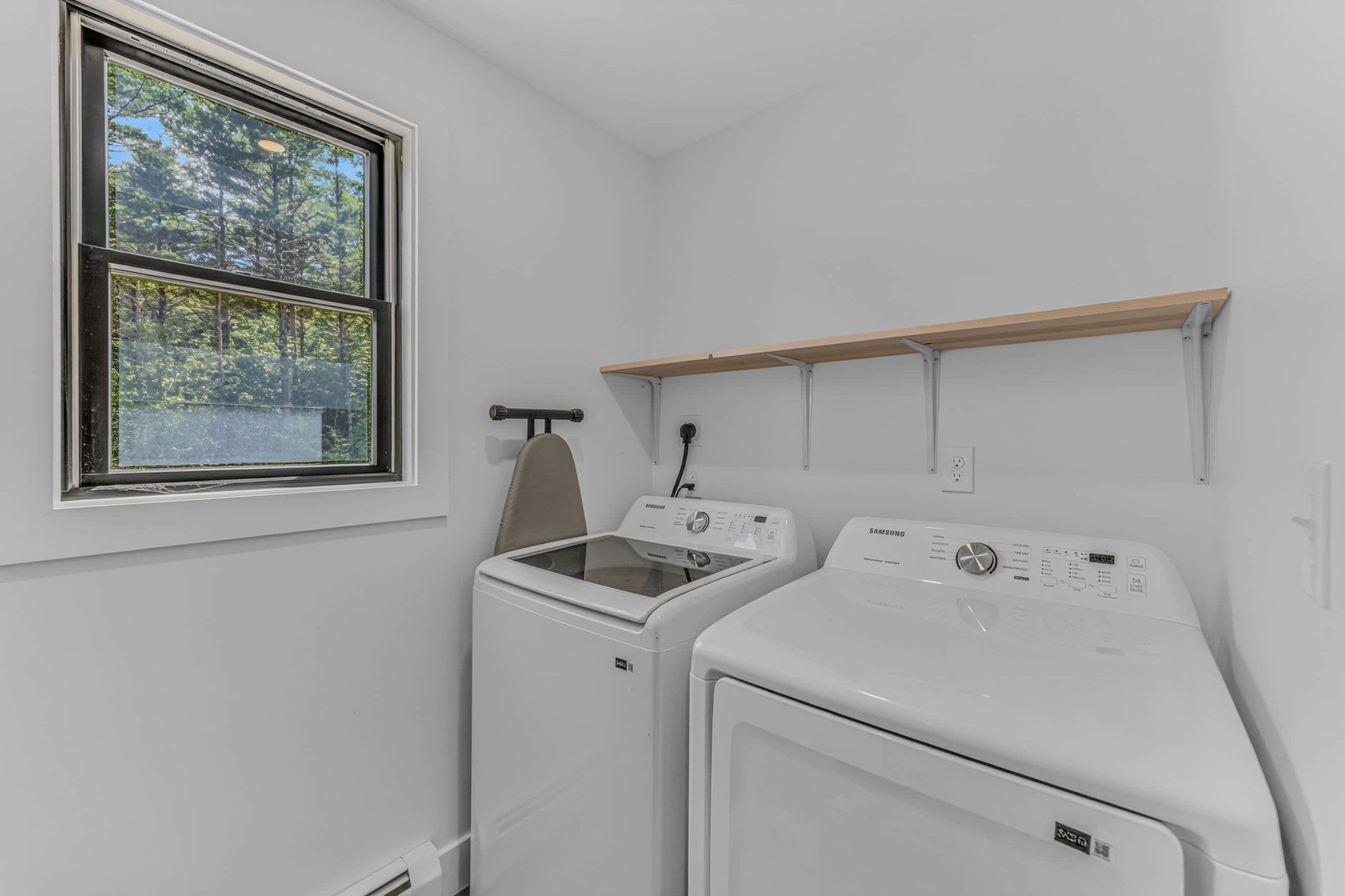 A laundry room with a window, a washing machine, a dryer, and a wooden shelf.
