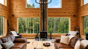 Interior acogedor de una cabaña con paredes de madera, chimenea, sofás de cuero y ventanas amplias con vistas al bosque.