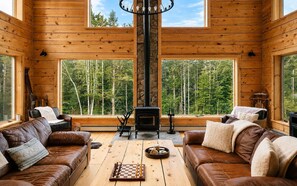 A cozy cabin interior with wooden walls, a fireplace, leather sofas, and large windows offering a forest view. - The Grafton Lodge Secluded Green Mountain Escape (Grafton)