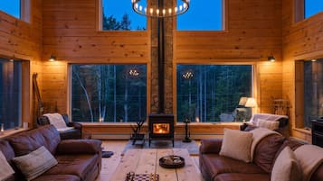 A cozy cabin interior with wooden walls, a fireplace, leather sofas, and large windows overlooking a forest.