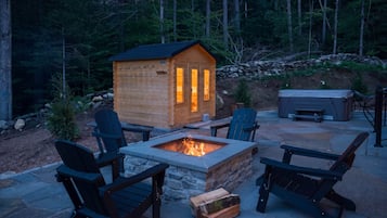 A wooden cabin with a fire pit in the center, surrounded by Adirondack chairs and a stone fire table.