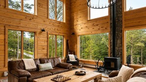 Interior acogedor de una cabaña con paredes de madera, ventanas amplias, chimenea y un tablero de ajedrez sobre una mesa.