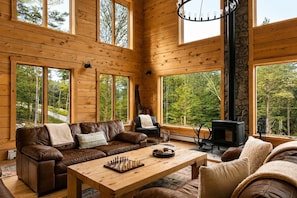A cozy cabin interior with wooden walls, large windows, a fireplace, and a chessboard on a table. - The Grafton Lodge Secluded Green Mountain Escape (Grafton)