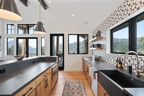 A modern kitchen with a large island, black countertops, wooden cabinets, and a view of the mountains through large windows. - Bear Scratch Mountain Views Modern Luxury (Crested Butte)
