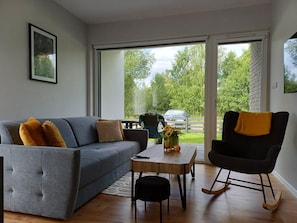 Een moderne woonkamer met een grijze bank, een zwarte schommelstoel, een houten salontafel en een uitzicht door grote glazen deuren op het buitengebied.