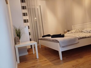 Un dormitorio con cama blanca, una mesita de noche, un banco, una planta en maceta y una ventana con persianas verticales.