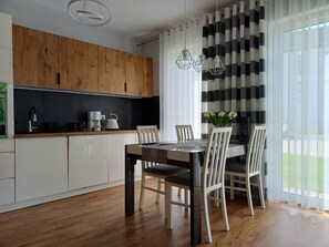 Een moderne keuken met houten kasten, een eettafel met stoelen en uitzicht op het groen door dunne gordijnen.
