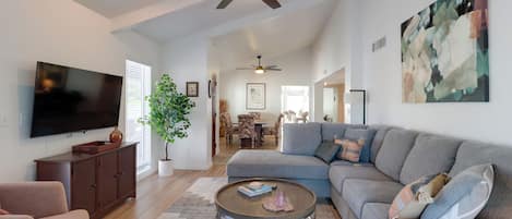 A spacious living room with a large grey sectional sofa, a flat-screen TV mounted on the wall, a ceiling fan, and a modern abstract painting.