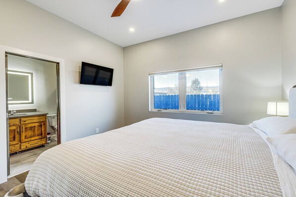 A bedroom with a large bed, a television mounted on the wall, a window with a view of a blue fence, and a wooden dresser.