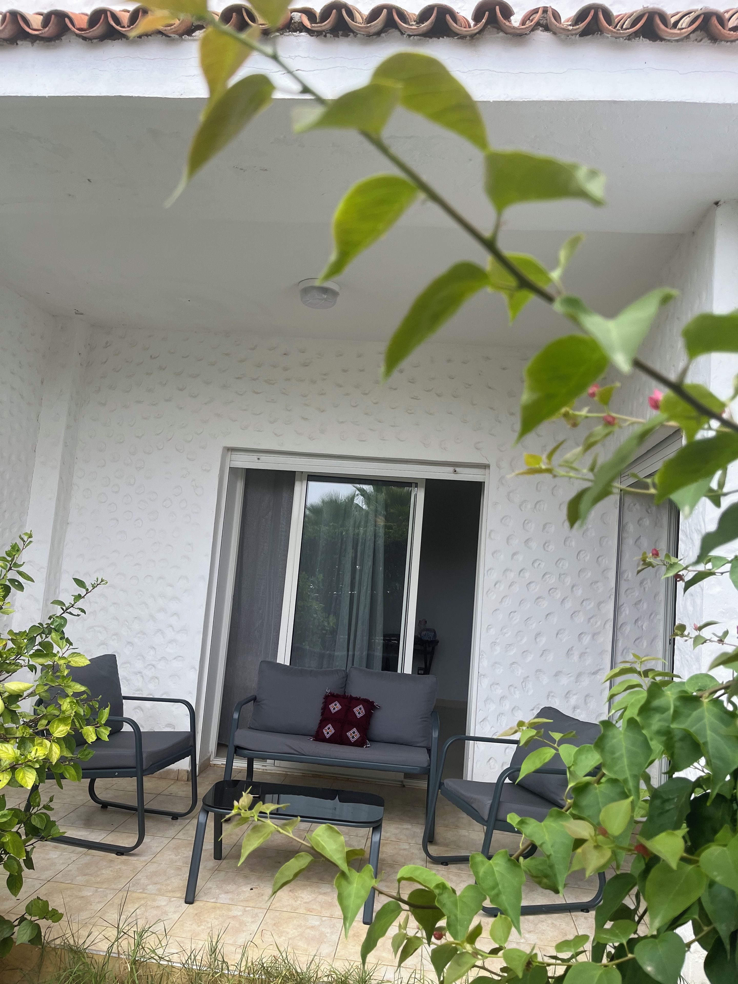 A covered outdoor seating area with a sofa and chairs, a small table, and a sliding glass door leading to an interior space.