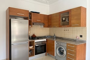 A compact kitchen with wooden cabinets, a stainless steel refrigerator, an oven, a washing machine, and a countertop with a sink.