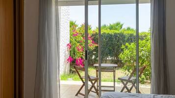 A room with a bed, a sliding glass door leading to an outdoor area with a table and chairs, and a view of greenery and flowers.