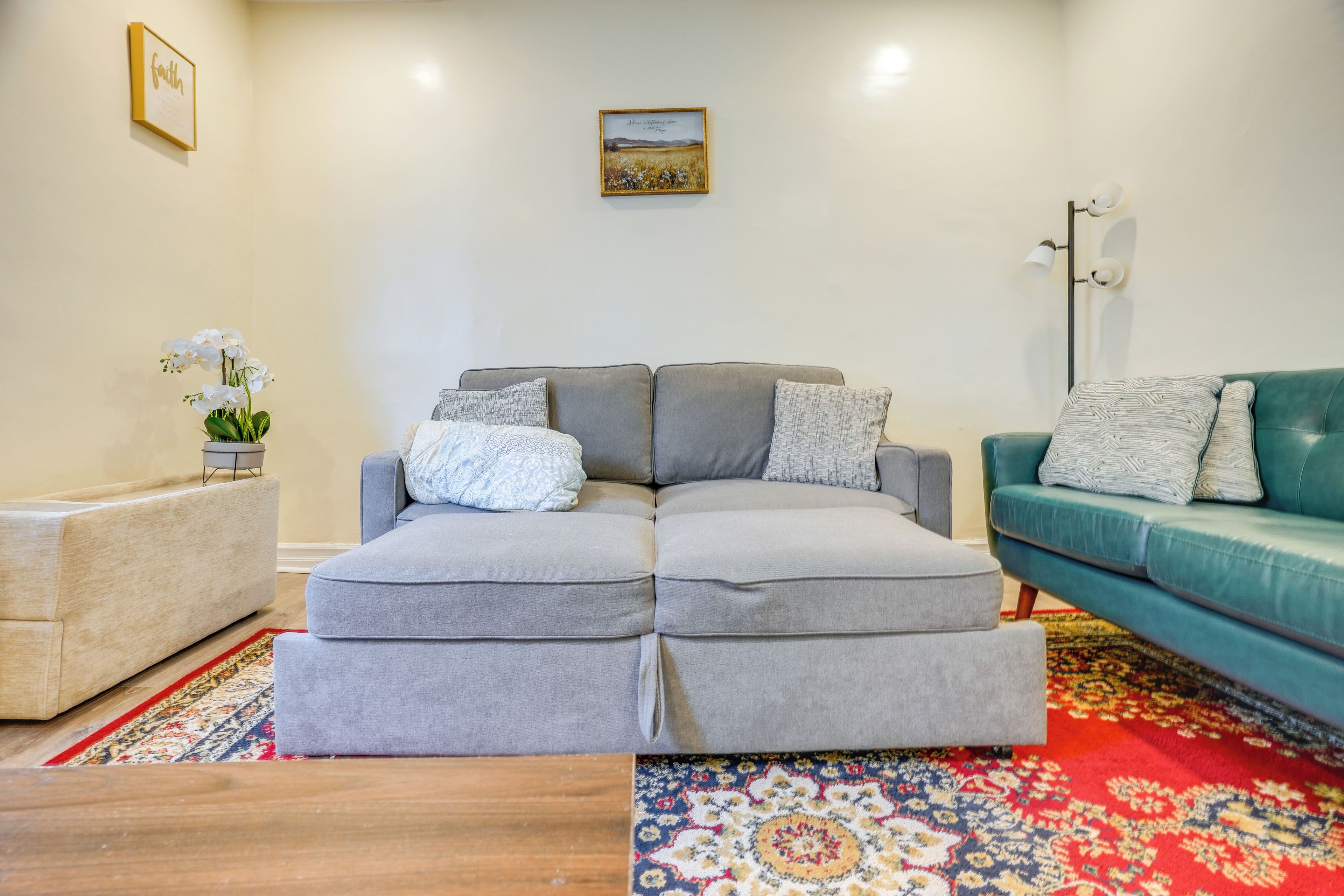 Una sala de estar con un sofá seccional gris, un sillón verde azulado y una mesita. Hay una fotografía enmarcada en la pared y un florero con flores blancas sobre una mesita auxiliar.