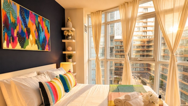Een hotelkamer met een groot bed, een kleurrijk schilderij, uitzicht op de stad en een dienblad met handdoeken en toiletartikelen.