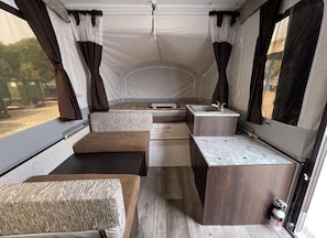 A compact interior with a kitchenette, a seating area, and a window providing a view of the outside. - Jayco Light Pop Up Trailer (Ajax)