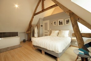 Un dormitorio con techo inclinado, una cama con ropa blanca, una silla de madera y una mesita de noche pequeña.
