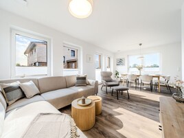 Una sala moderna con un sofá, sillones, un comedor con mesa y sillas, y ventanas amplias que ofrecen vistas a un edificio.