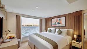 A hotel room with a large bed, a desk with a fruit bowl, a bedside table with a lamp, and a view of the water. - MS Royal Serenade (Luxor)