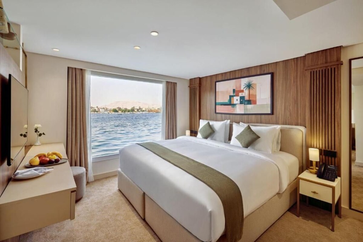 Habitación de hotel con una cama grande, un escritorio con frutero, una mesita de noche con lámpara y vista al agua.