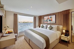 Habitación de hotel con una cama grande, un escritorio con frutero, una mesita de noche con lámpara y vista al agua.