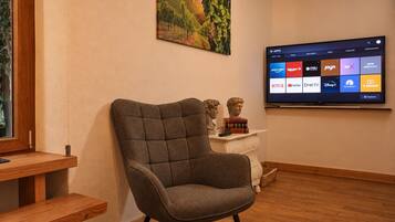 Een woonkamer met een grijze fauteuil, een houten salontafel, een flatscreen-tv waarop een streamingdienst draait, en een schilderij aan de muur.