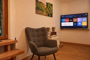 Een woonkamer met een grijze fauteuil, een houten salontafel, een flatscreen-tv waarop een streamingdienst draait, en een schilderij aan de muur.