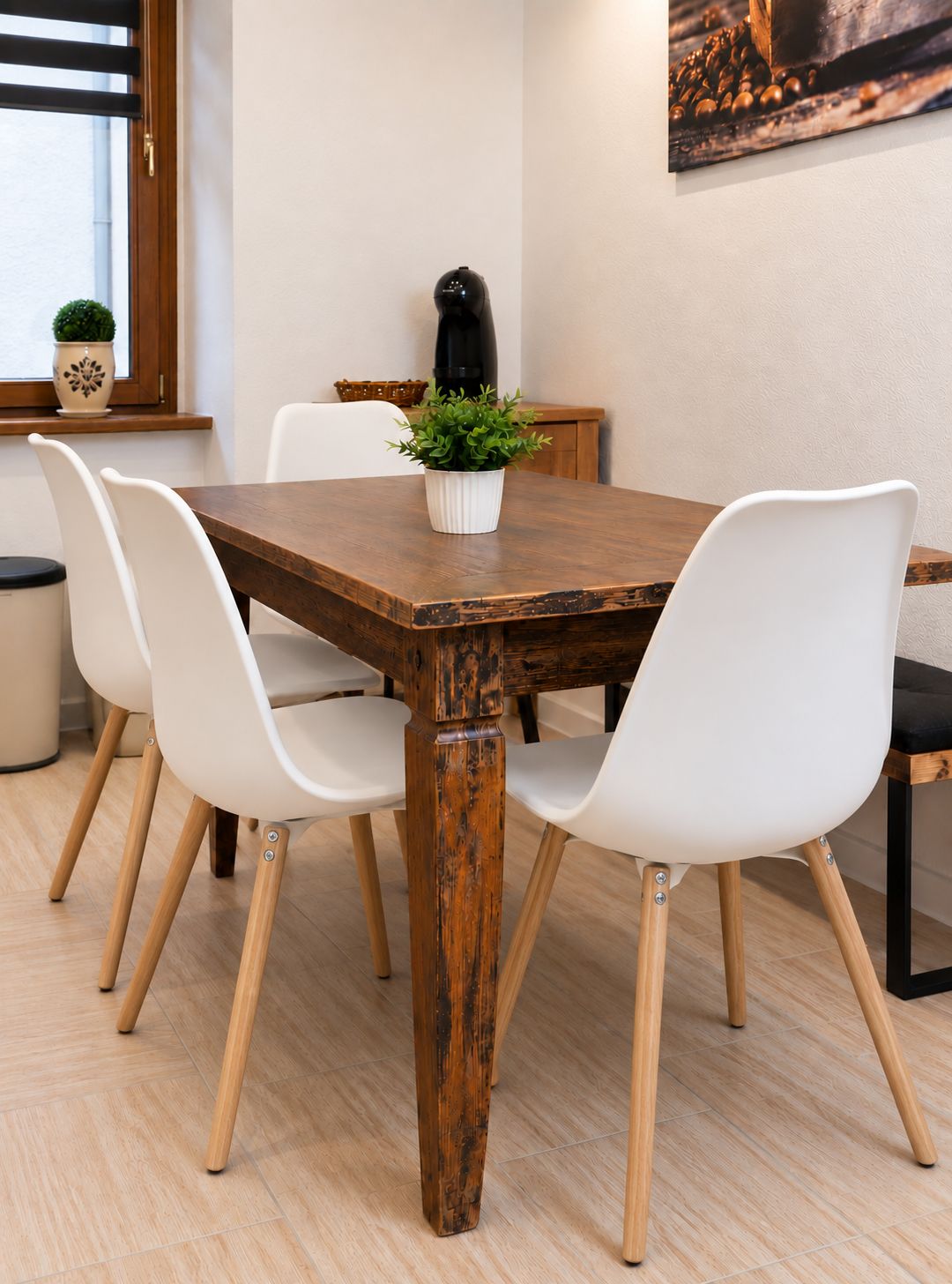 Een eetgedeelte met een houten tafel, witte stoelen, een potplant en een koffieapparaat.